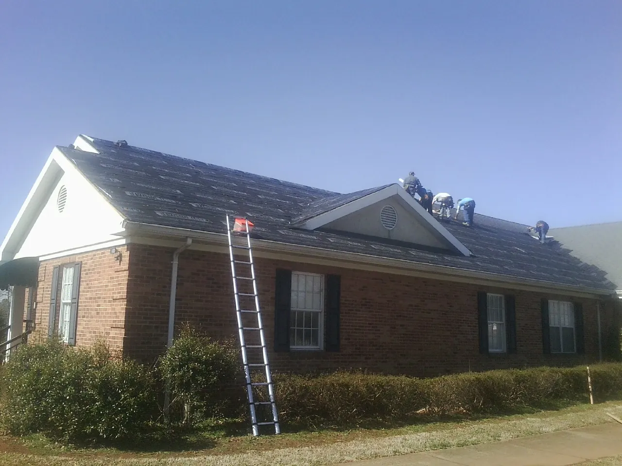 Crew tearing shingles off a brick ranch roof with a ladder set — Emergency Roof Repair work by our crew in Whitsett