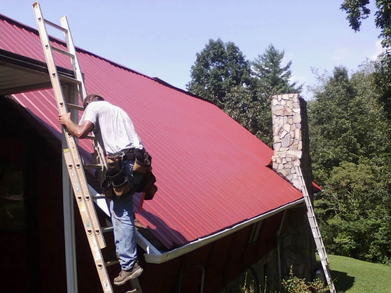 Roofer on a ladder beside a red metal roof and stone chimney — Soffit & Fascia Repair by Roofing Brothers in Whitsett